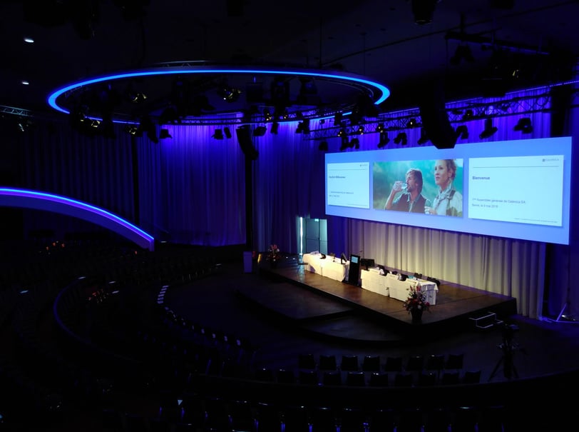 Large auditorium with illuminated stage on which a podium stands and a large screen with three slides is displayed above it